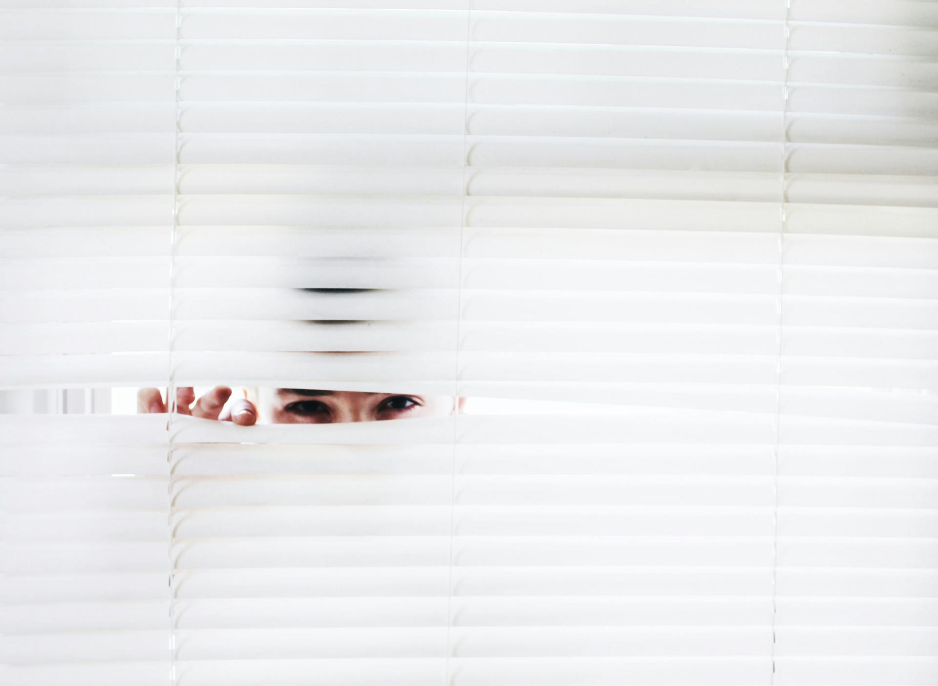 Person peers through window blinds into restricted area; concept illustration of privacy inspection.